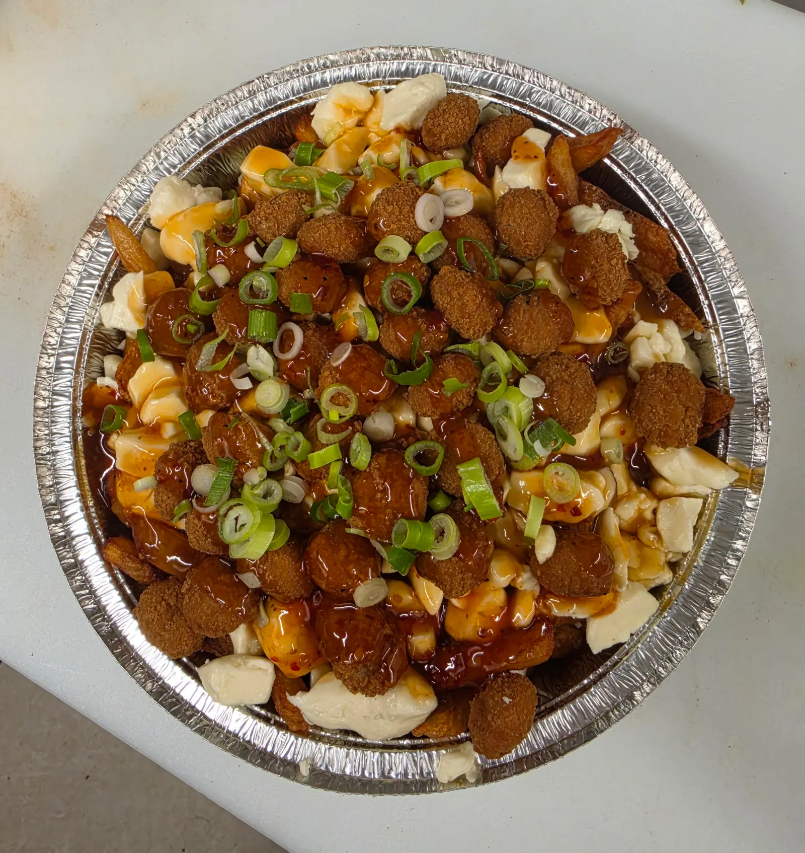 Poutine garnie de bouchées panées, de sauce luisante et d'oignon vert, dans une barquette ronde en aluminium