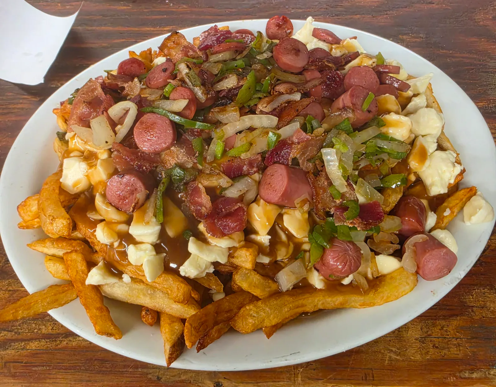 Poutine garnie de rondelles de saucisse, de bacon, d'oignons et de poivron vert, dans une assiette blanche
